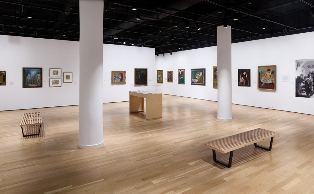 Art gallery with light wooden floors, framed paintings and a blown-up photo reproduction on white walls, a wooden bench, and a central display case.
