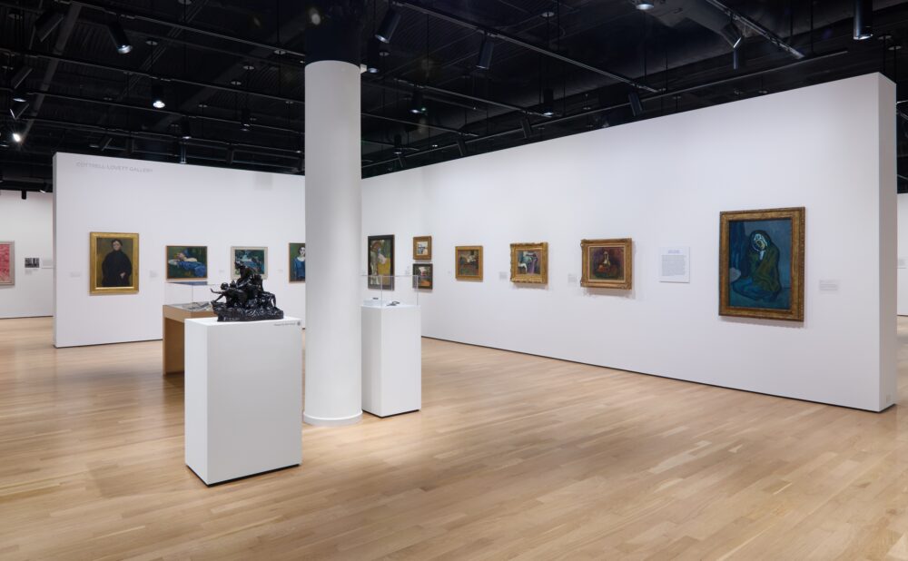 Art gallery with framed paintings and sculptures on white walls, featuring a prominent white column and wooden flooring.
