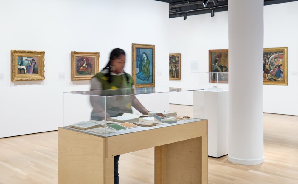 Modern art gallery with white walls and a column, with framed turn-of-the-century European paintings on the wall; at center, a Black woman examines items in a display case.