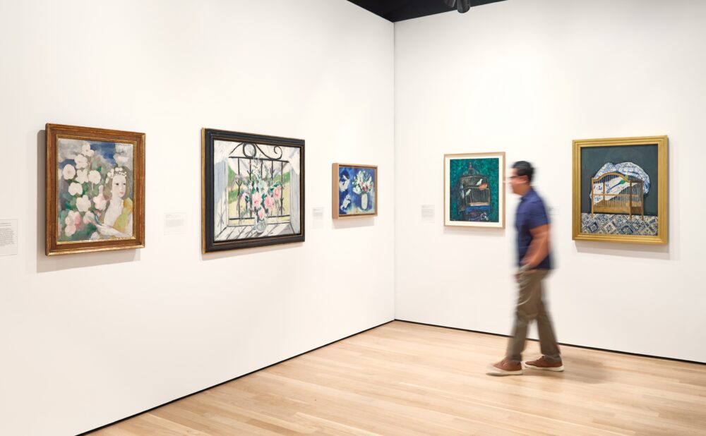 Corner of a modern gallery with framed paintings hung nearly on the wall, while a man in a blue shirt and beige pants walks by with his hands in his pockets
