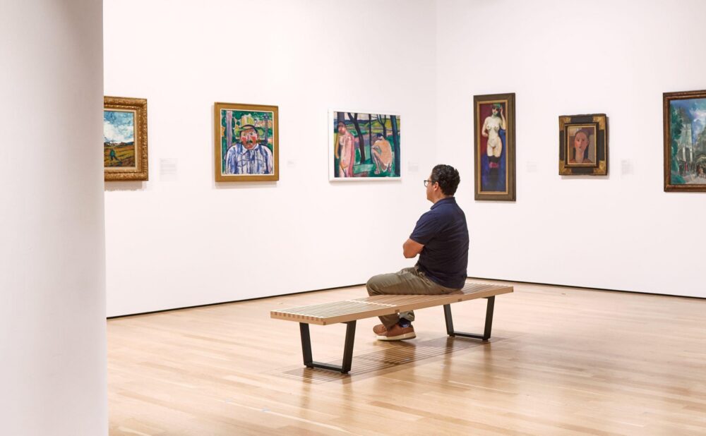 A person sits on a bench in an art gallery looking at several paintings on a white wall.