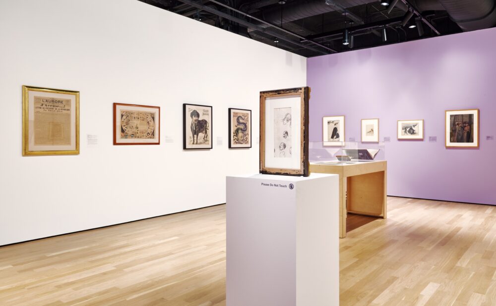 Modern art gallery with white and lavendar walls and a light wood floor. Framed illustrations and drawings are hung on the wall, with one displayed on a centrally placed pedestal. A display case is visible in the background.