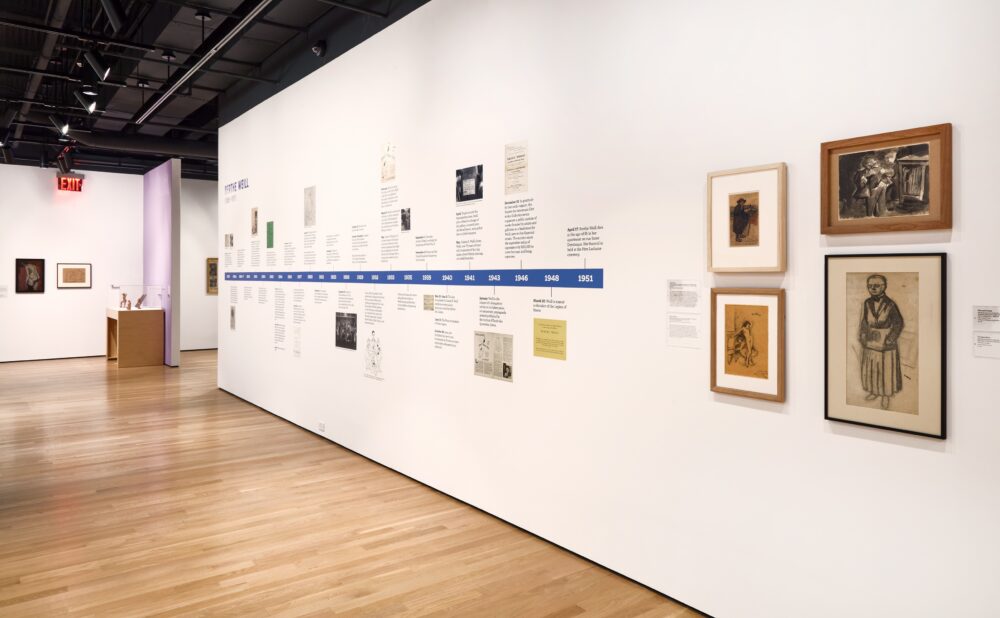 Art gallery with white walls, wooden floors, framed artworks, and a timeline.
