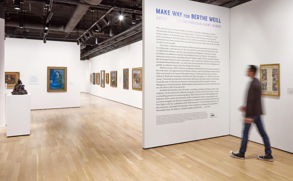 Modern art gallery with exhibition title and introductory text on a wall. A man walk towards the wall and into the museum, where framed paintings and sculptures can be seen in the white-walled galleries.