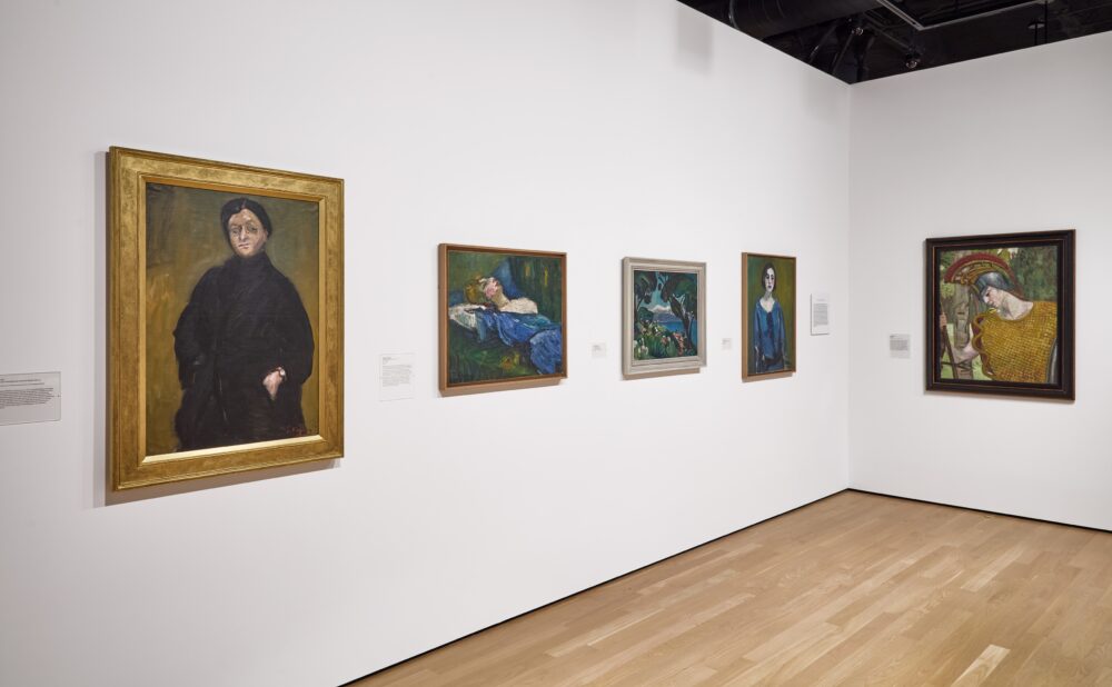 Art gallery with framed paintings displayed on a white wall with a light wooden floor.