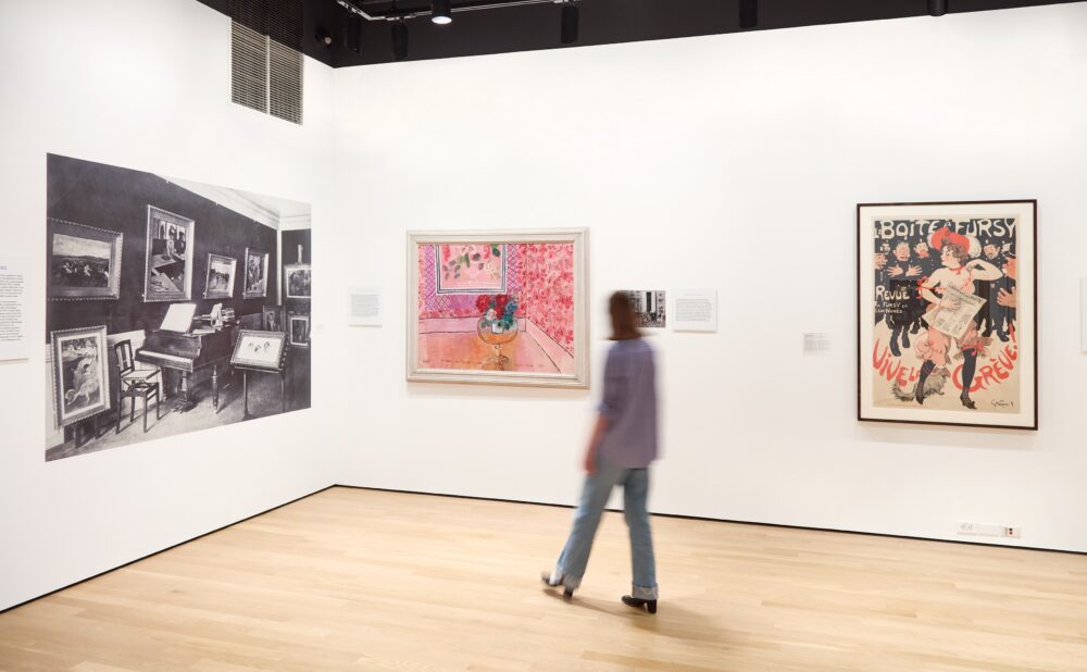Art gallery with a mural, painting, and vintage poster, with a person walking through.