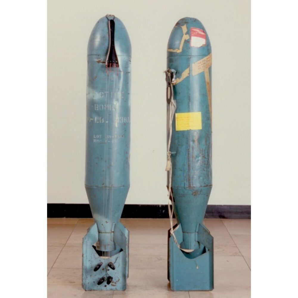 Two tall, bomb-shaped metal casings standing vertically side-by-side in a gallery setting. (Alt-text generated by AI)