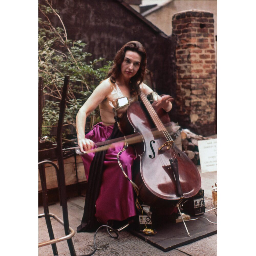 Color photograph of Charlotte Moorman playing a cello outdoors in front of a brick wall. (Alt-text generated by AI)