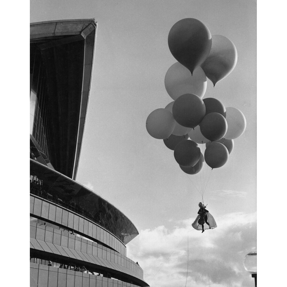Black-and-white aerial performance photograph showing Charlotte Moorman suspended in mid-air beneath a cluster of large balloons during "Sky Kiss". (Alt-text generated by AI)