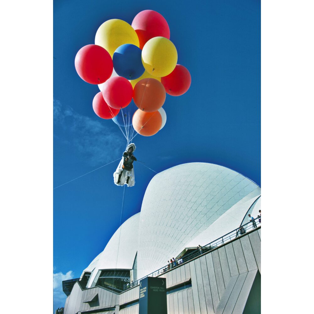 Vibrant color photograph of Charlotte Moorman floating in the sky above the Sydney Opera House tied to a bundle of colorful balloons during her "Sky Kiss" performance. (Alt-text generated by AI)