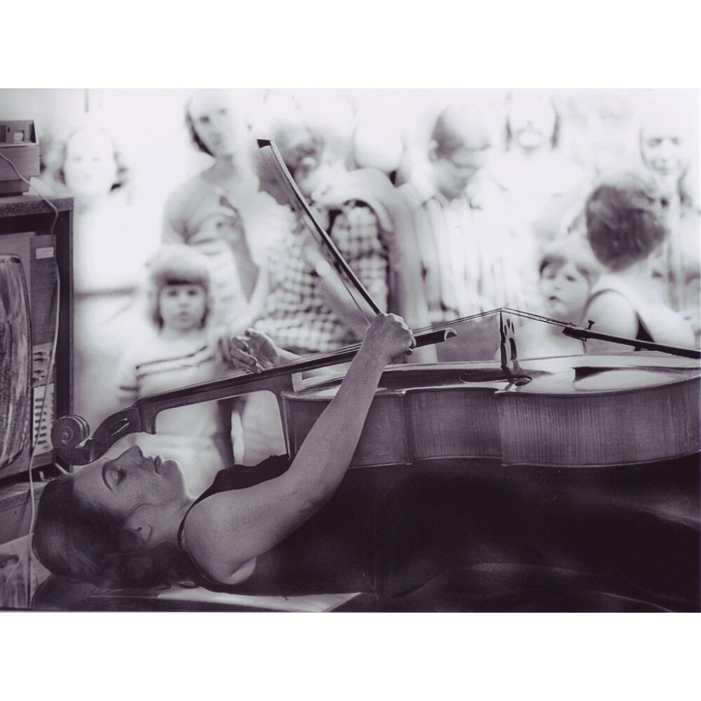 Black-and-white close-up photograph of Charlotte Moorman lying down while playing a cello suspended over her while adults and children watch. (Alt-text generated by AI)