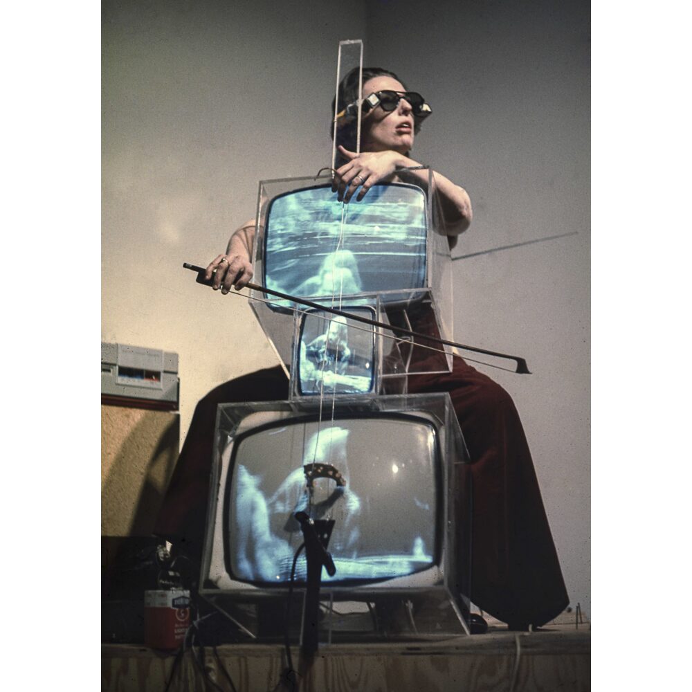 Performance photo showing Charlotte Moorman playing the "TV Cello" composed of stacked, illuminated television monitors. (Alt-text generated by AI)