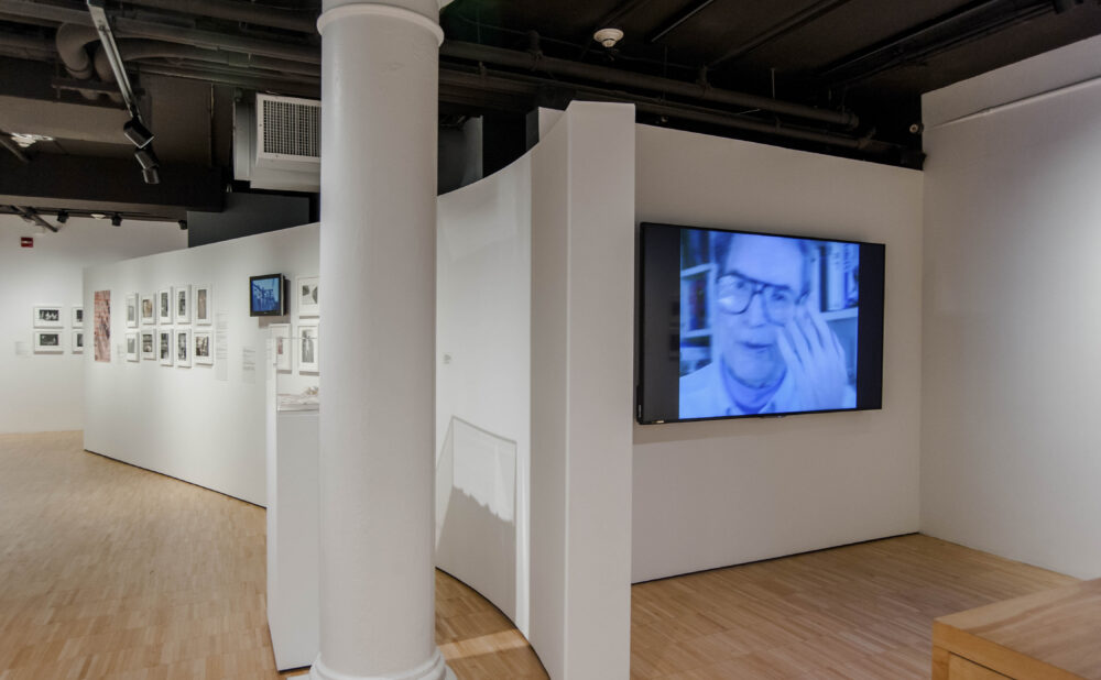 Gallery space centered around a large white pillar, with a video monitor mounted on a partition wall on the right. (Alt-text generated by AI)