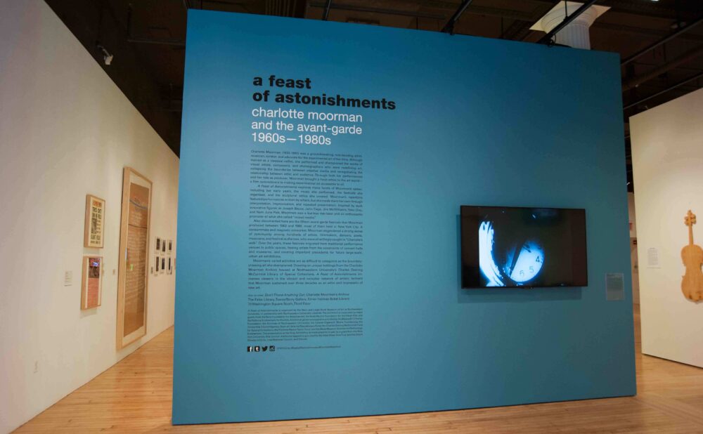 Exhibition entry featuring a large teal accent wall mounted with introductory wall text and a video monitor. (Alt-text generated by AI)