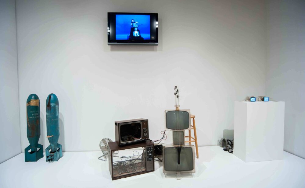 Exhibition display featuring two bomb-shaped casings, video monitors, and Nam June Paik's "TV Cello" against a white wall. (Alt-text generated by AI)