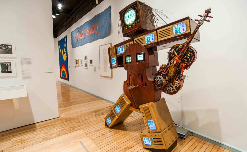 Gallery space showcasing Nam June Paik's "K-456" robot sculpture standing near a white wall with surrounding artworks. (Alt-text generated by AI)