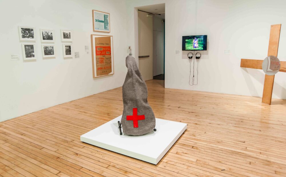 View of a gallery room featuring a central pedestal sculpture wrapped in cloth with a red cross, flanked by wall-mounted artworks. (Alt-text generated by AI)