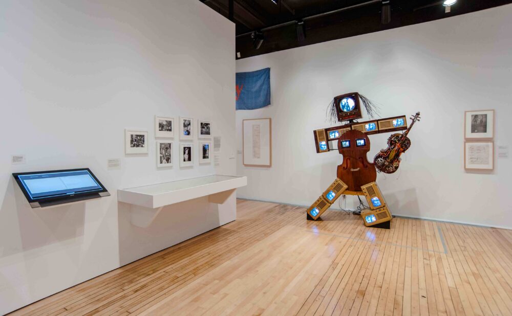 Wide view of the gallery interior displaying free-standing sculptures, small wall-mounted photographs, and interactive touchscreens. (Alt-text generated by AI)