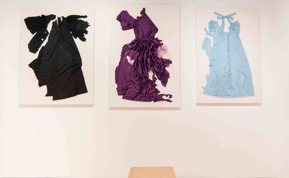 White gallery wall featuring three tattered dresses in black, purple, and light blue, eachmounted on a canvas. (Alt-text generated by AI)