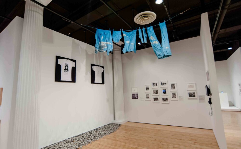 Exhibition space featuring garments suspended from the ceiling and framed photographs mounted on surrounding white walls. (Alt-text generated by AI)