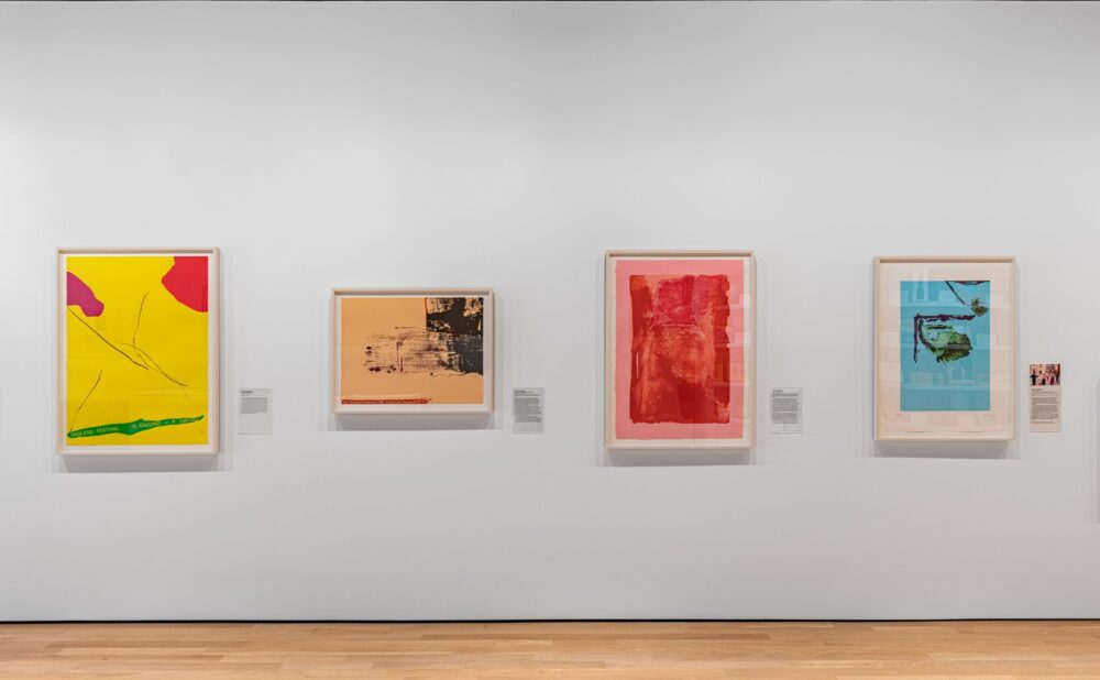 A museum gallery wall displays four framed artworks arranged horizontally on a white wall above a pale wood floor. Each artwork has a small wall label positioned nearby. Alt text generated by AI.