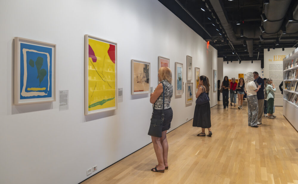 Visitors view framed artwork in a bright contemporary gallery with a bookstore shelf along the right wall.