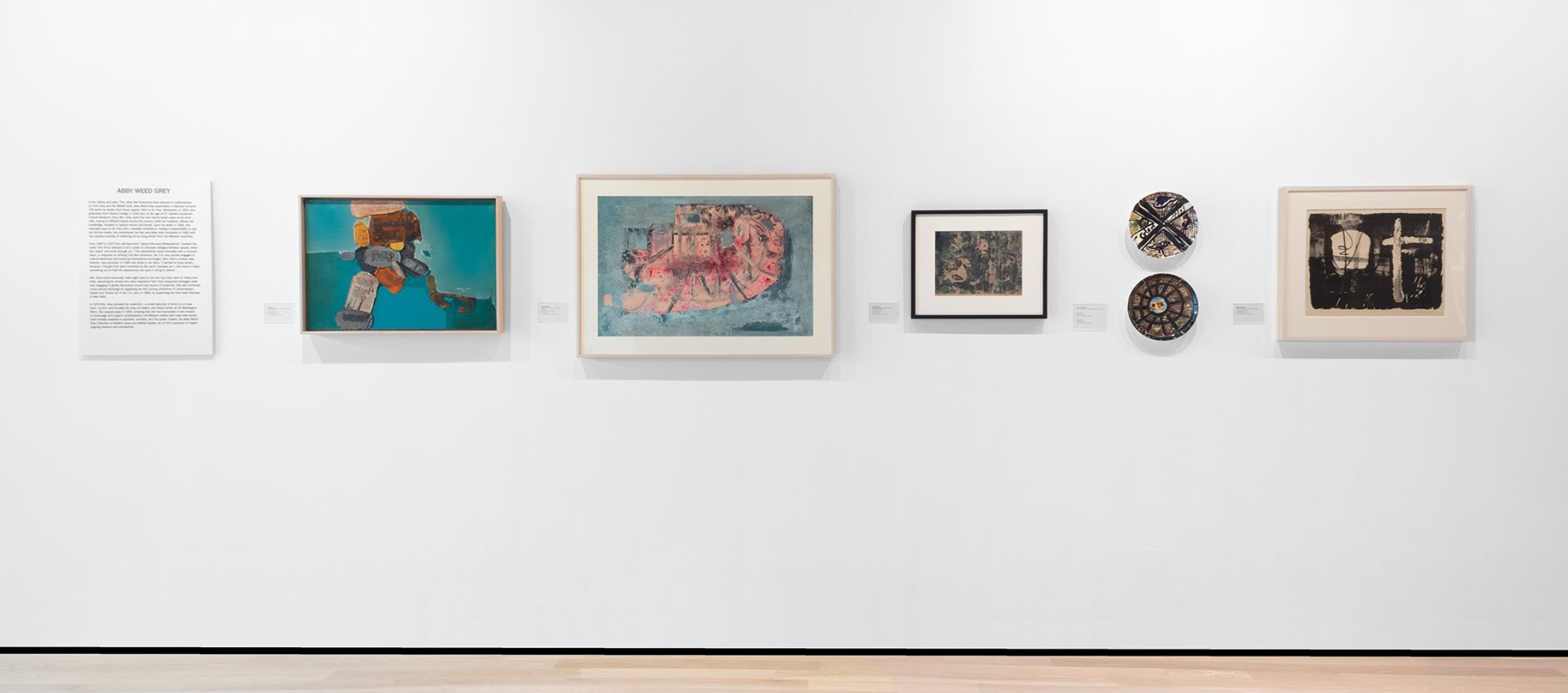 A gallery wall displaying six framed contemporary artworks, including paintings and ceramics, and a text panel.