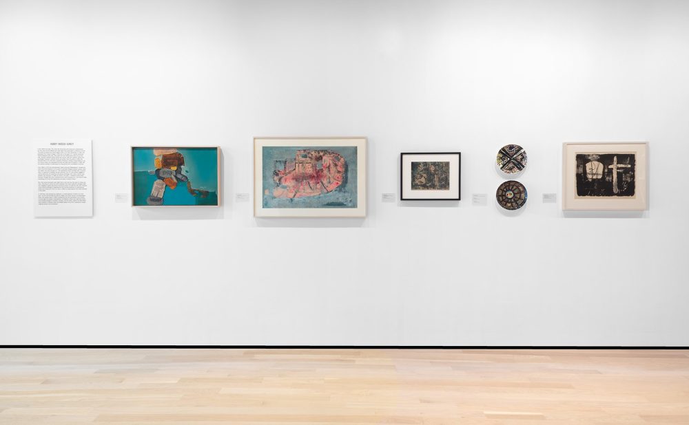 A gallery wall displaying six framed contemporary artworks, including paintings and ceramics, and a text panel.