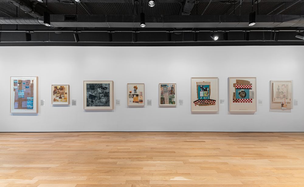 Art gallery with framed artworks on a white wall, wooden floor, and black industrial ceiling.