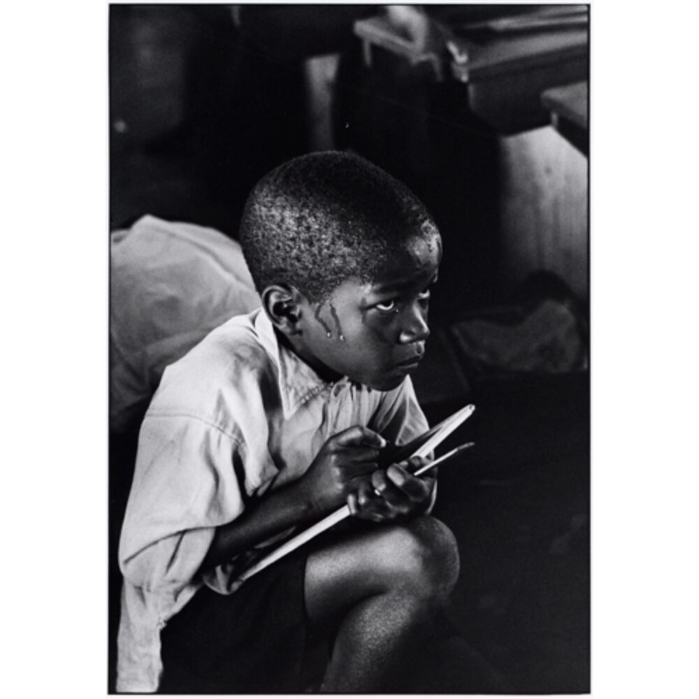 Black-and-white photograph of a young boy crouched down holding a slate board and writing utensil, looking off-camera and visibly sweating. (Alt-text generated by AI)