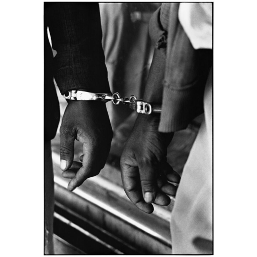 High-contrast close-up photograph of handcuffed hands belonging to two individuals standing side-by-side. (Alt-text generated by AI)