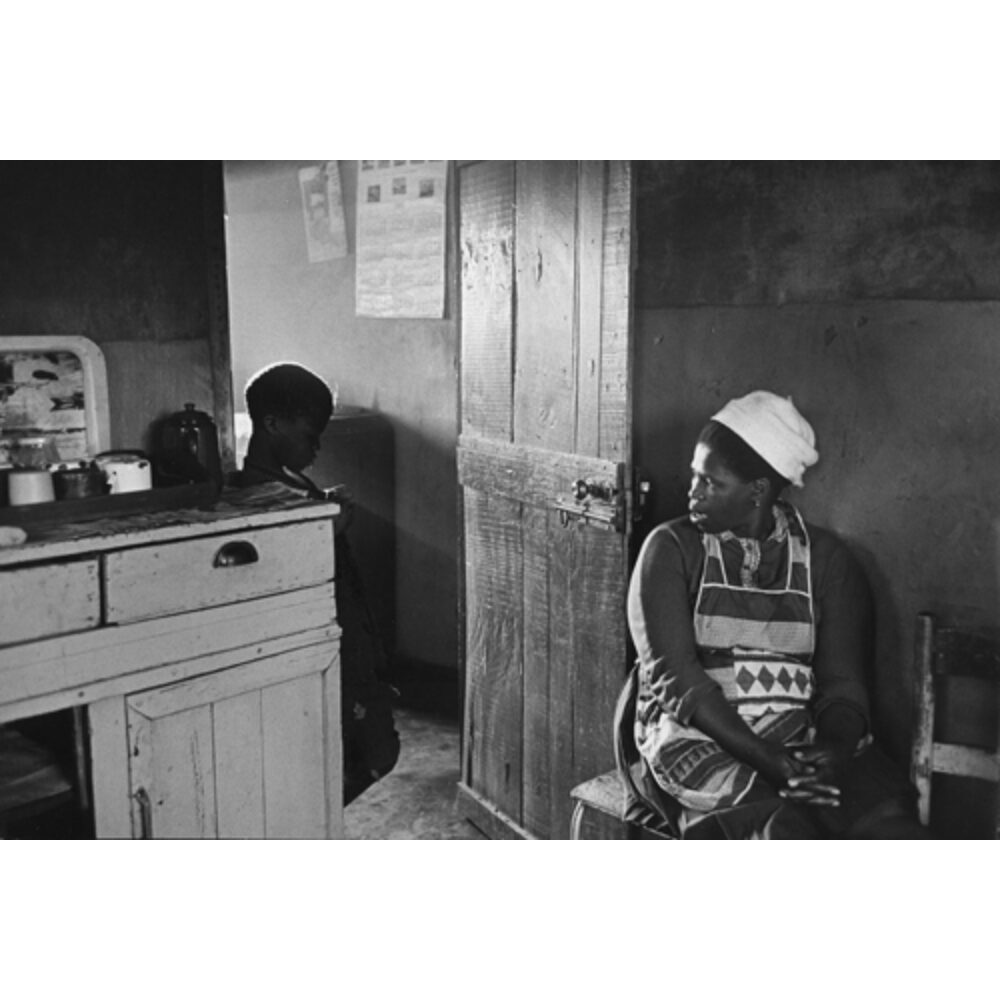 Interior black-and-white photograph depicting a woman seated near an open doorway while a child looks on from inside the room. (Alt-text generated by AI)
