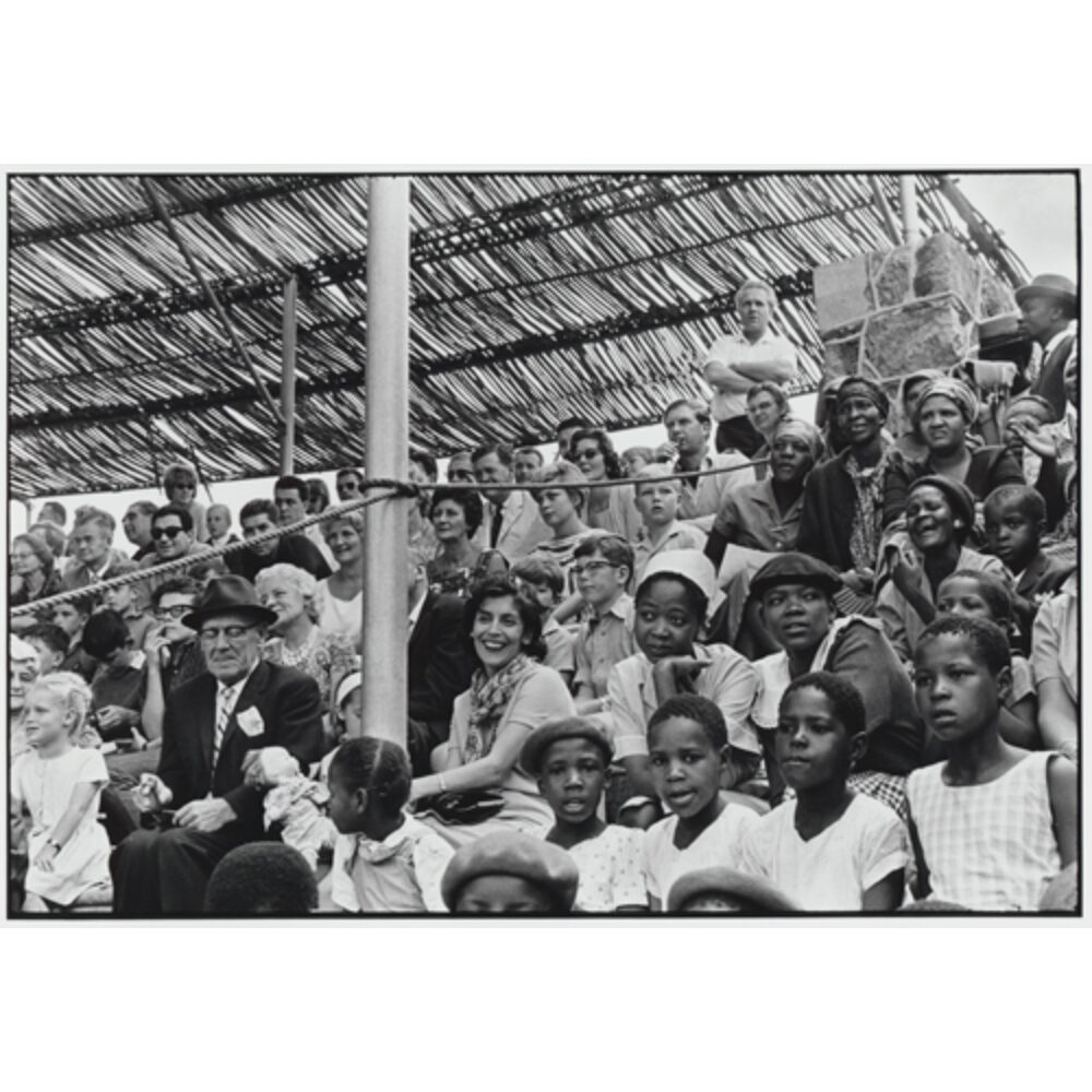 Crowd photograph showing spectators seated together under a thatched roof structure at an outdoor event. (Alt-text generated by AI)