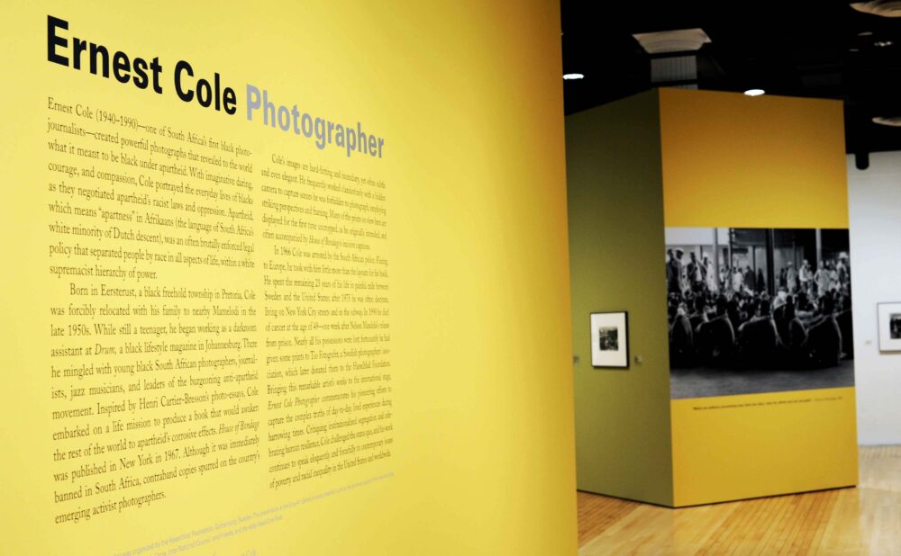 Exhibition entrance featuring a bold yellow wall printed with biographical text titled "Ernest Cole Photographer" alongside a large black-and-white photo enlargement. (Alt-text generated by AI)