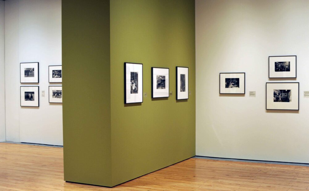 Gallery interior view showing muted green partition walls hung with framed black-and-white documentary photographs above light hardwood floors. (Alt-text generated by AI)