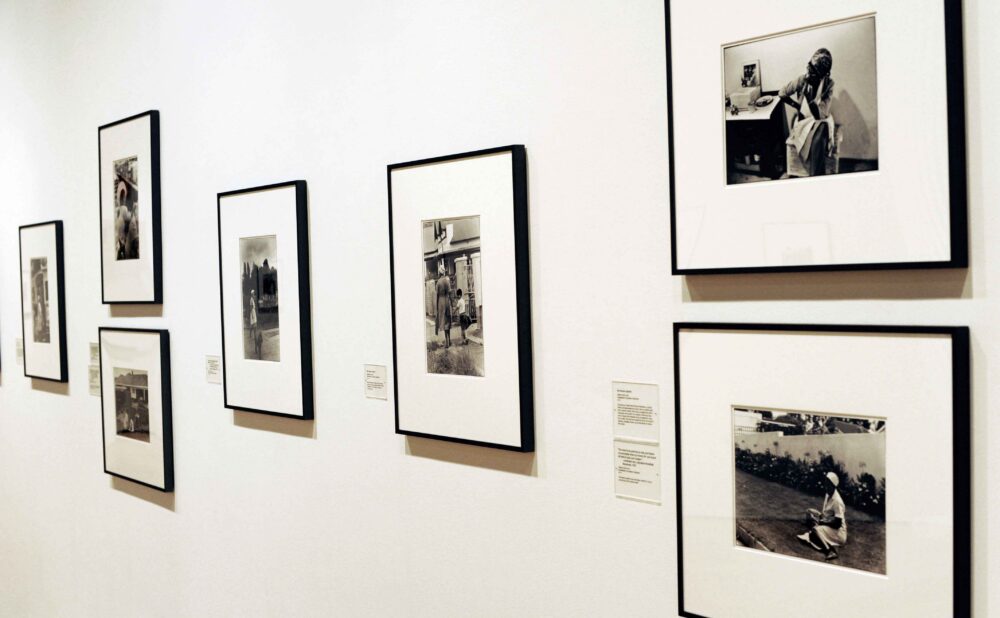 Perspective view along a gallery wall displaying a row of black-and-white photographs mounted in black frames with white matting, each accompanied by small wall labels. (Alt-text generated by AI)