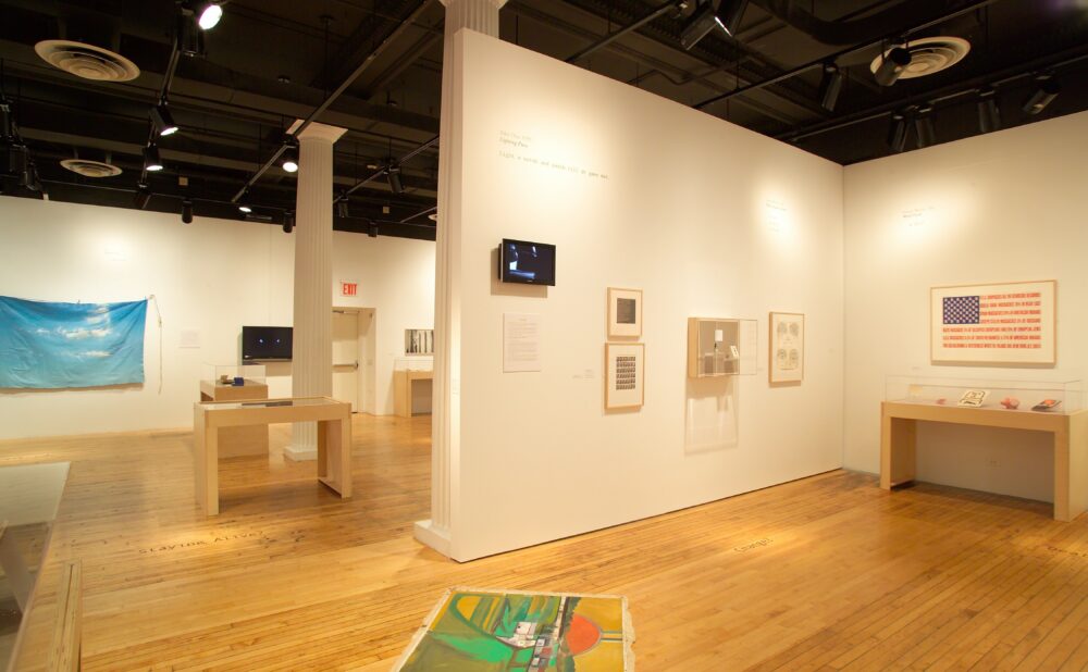 An art gallery interior with white partition walls, wooden flooring, display tables, and various framed artworks and banners mounted on the walls. Alt text generated by AI