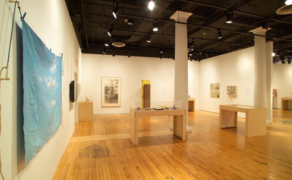 A wide shot of a gallery room with hardwood floors, a large blue banner hanging on the left wall, and multiple wood display tables positioned throughout the space. Alt text generated by AI