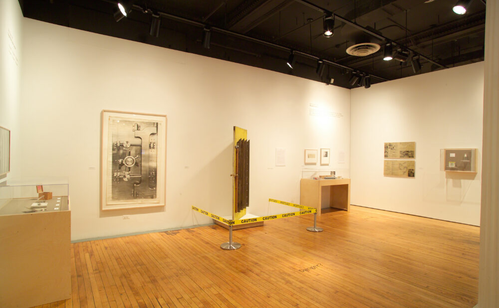 A gallery corner featuring framed art on white walls, a dark free-standing panel secured with yellow stanchion tape, and a small display table. Alt text generated by AI