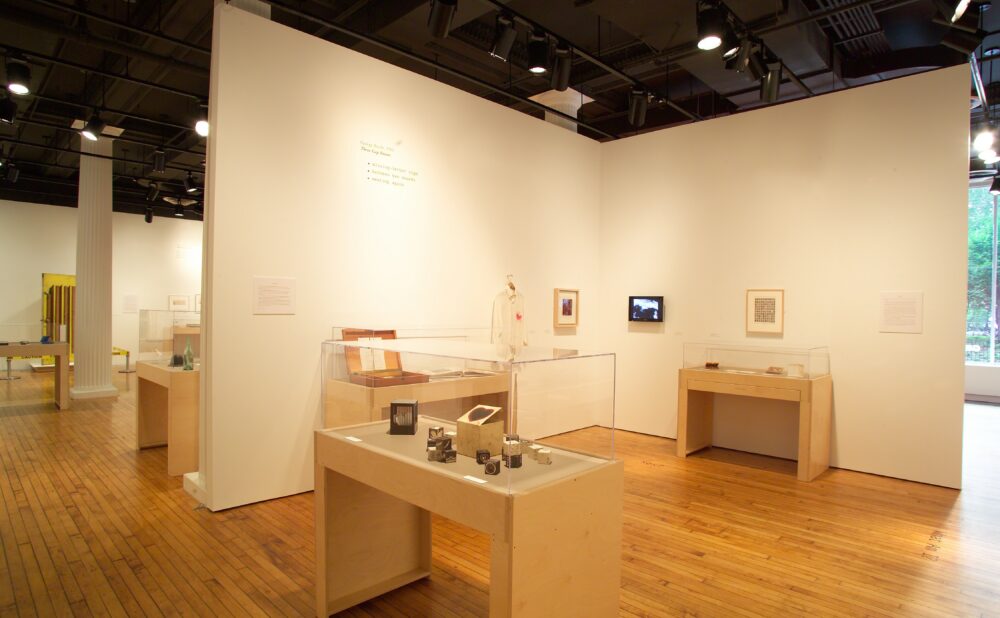 An exhibition space showcasing glass-topped wooden display cases containing small object collections, set against white gallery walls and wooden flooring. Alt text generated by AI