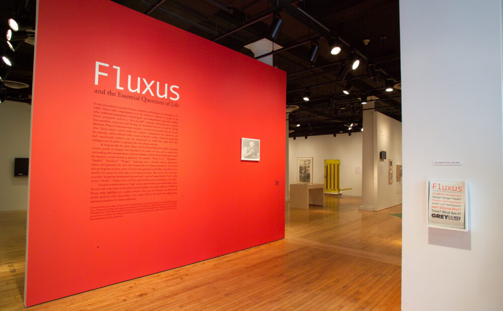 A prominent bright red exhibition wall titled "Fluxus" with intro text in an art gallery setting, leading toward other gallery sections. Alt text generated by AI
