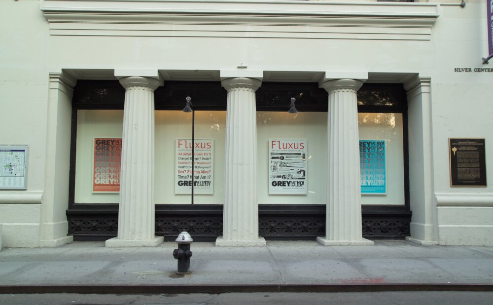 The exterior facade of a building featuring classical white columns, glass storefront windows displaying art posters, and street pillars. Alt text generated by AI