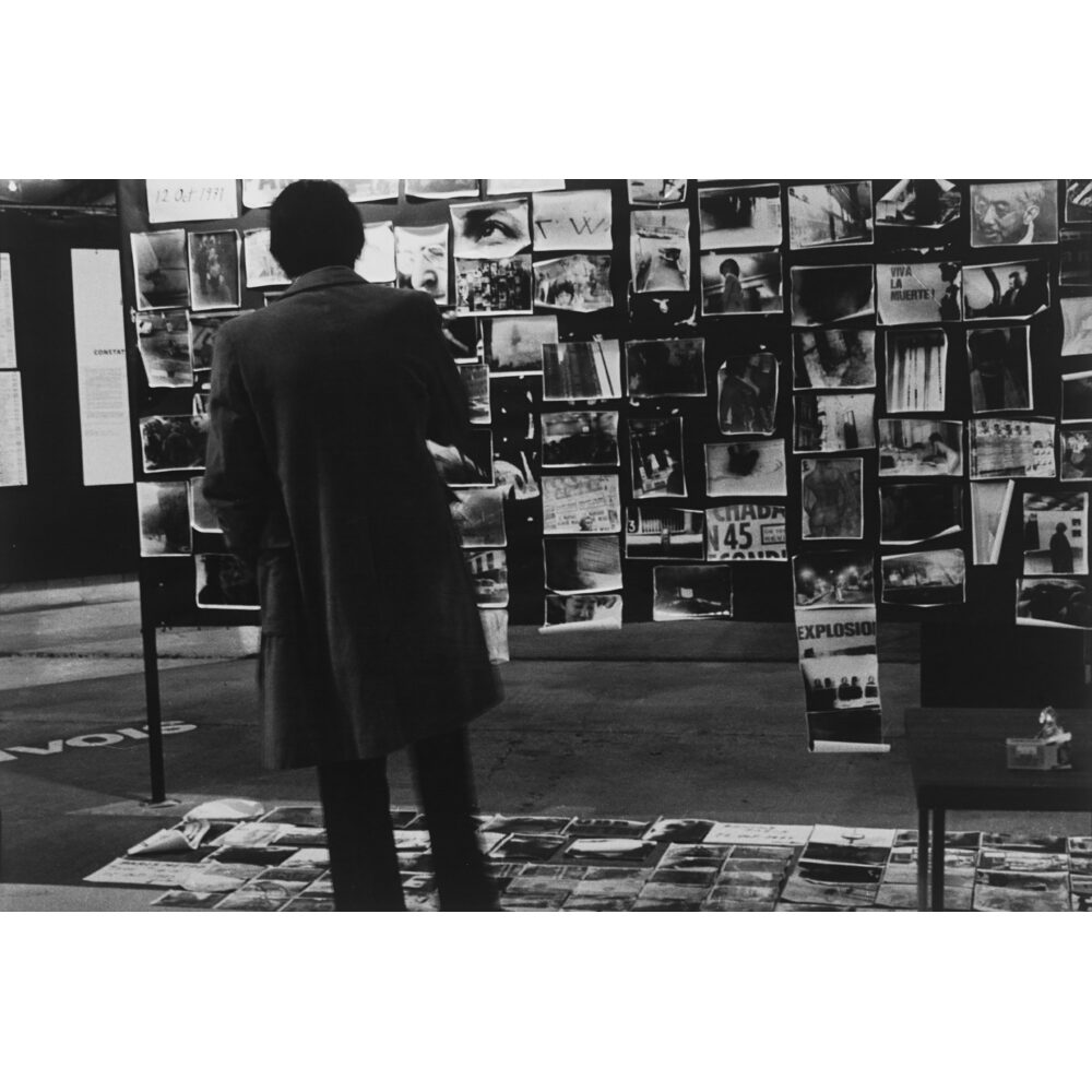 Documentary photograph by Takuma Nakahira showing a person standing in front of a grid wall covered in photographs. (Alt-text generated by AI)