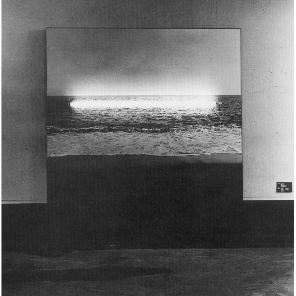 Black-and-white photograph showing a glowing horizontal tube light mounted above an ocean horizon scene by Masafumi Maita. (Alt-text generated by AI)