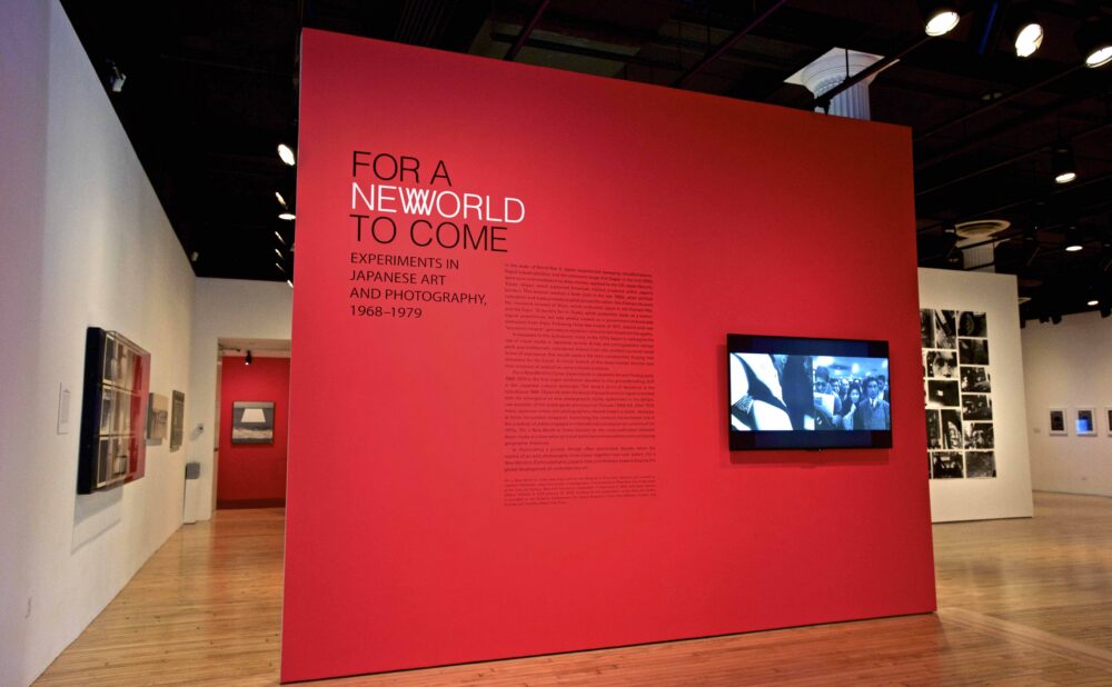 Exhibition entry view featuring a bright red accent wall with the title "FOR A NEW WORLD TO COME" and a wall-mounted video monitor. (Alt-text generated by AI)