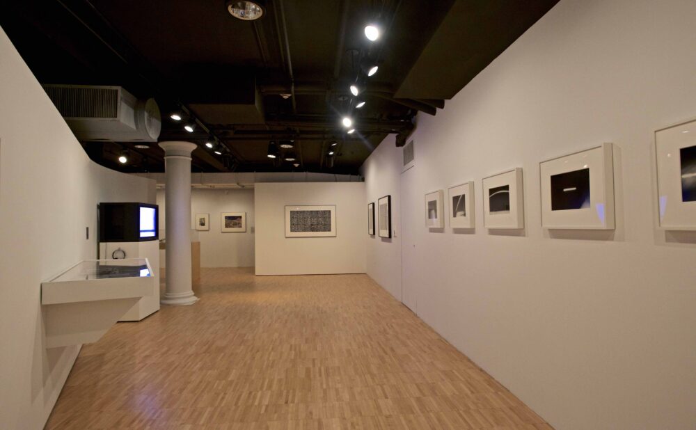 Perspective view down an exhibition hall featuring white walls hung with framed black-and-white photographs above hardwood floors. (Alt-text generated by AI)