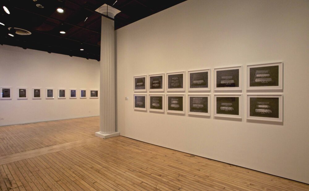 View of a white gallery wall mounted with an organized grid of twelve framed black-and-white photographs. (Alt-text generated by AI)