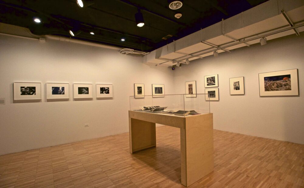 Corner exhibition space featuring a wooden vitrine flanked by framed photographic series on white walls. (Alt-text generated by AI)