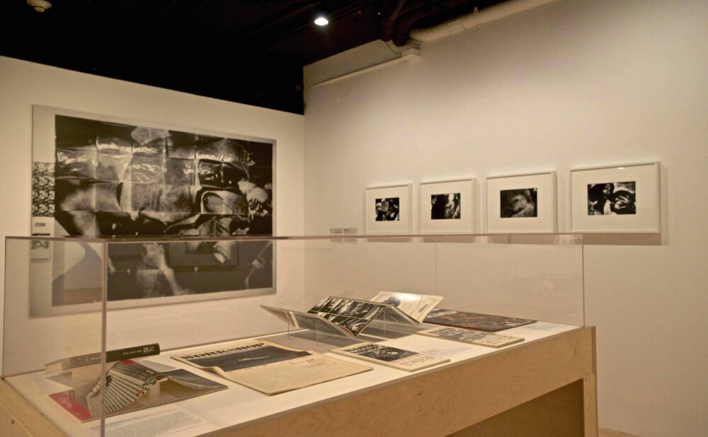 Wooden display vitrine under Plexiglas containing archival books and prints, backed by a large black-and-white photograph. (Alt-text generated by AI)