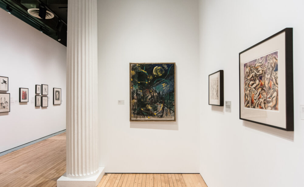 View down a gallery hallway featuring framed paintings on white walls alongside a structural pillar. (Alt-text generated by AI)